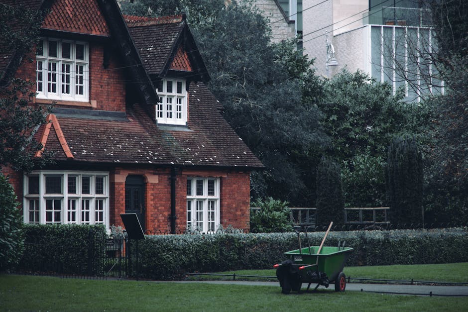 A well-maintained garden in front of a traditional red-brick house with white-framed windows, featuring a neatly trimmed hedge border along the front. The lawn is lush and green, with a pathway that r