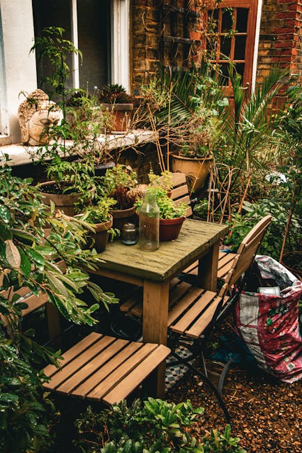 A cozy urban garden scene featuring a weathered wooden outdoor table surrounded by matching wooden benches and chairs, set against a brick wall with a wooden-framed window. The table is adorned with v